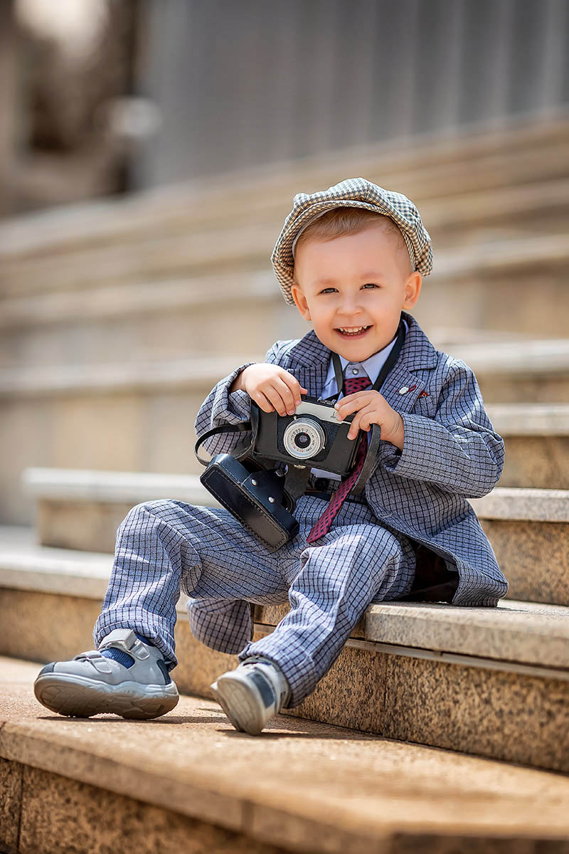Porträt eines kleinen jungen Fotografen, der mit einer Retro-Vintage-Fotokamera auf den Stufen draußen fotografiert. Fotoshooting für Kinder mit Mazelle Photography Fotostudio®