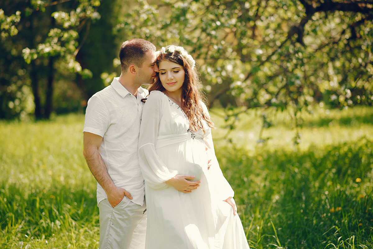 Schönes junges schwangeres Frau in einem langen weißen Kleid und Kranz auf dem Kopf, das mit ihrem Mann in einem sonnigen Sommerpark zwischen den Bäumen steht