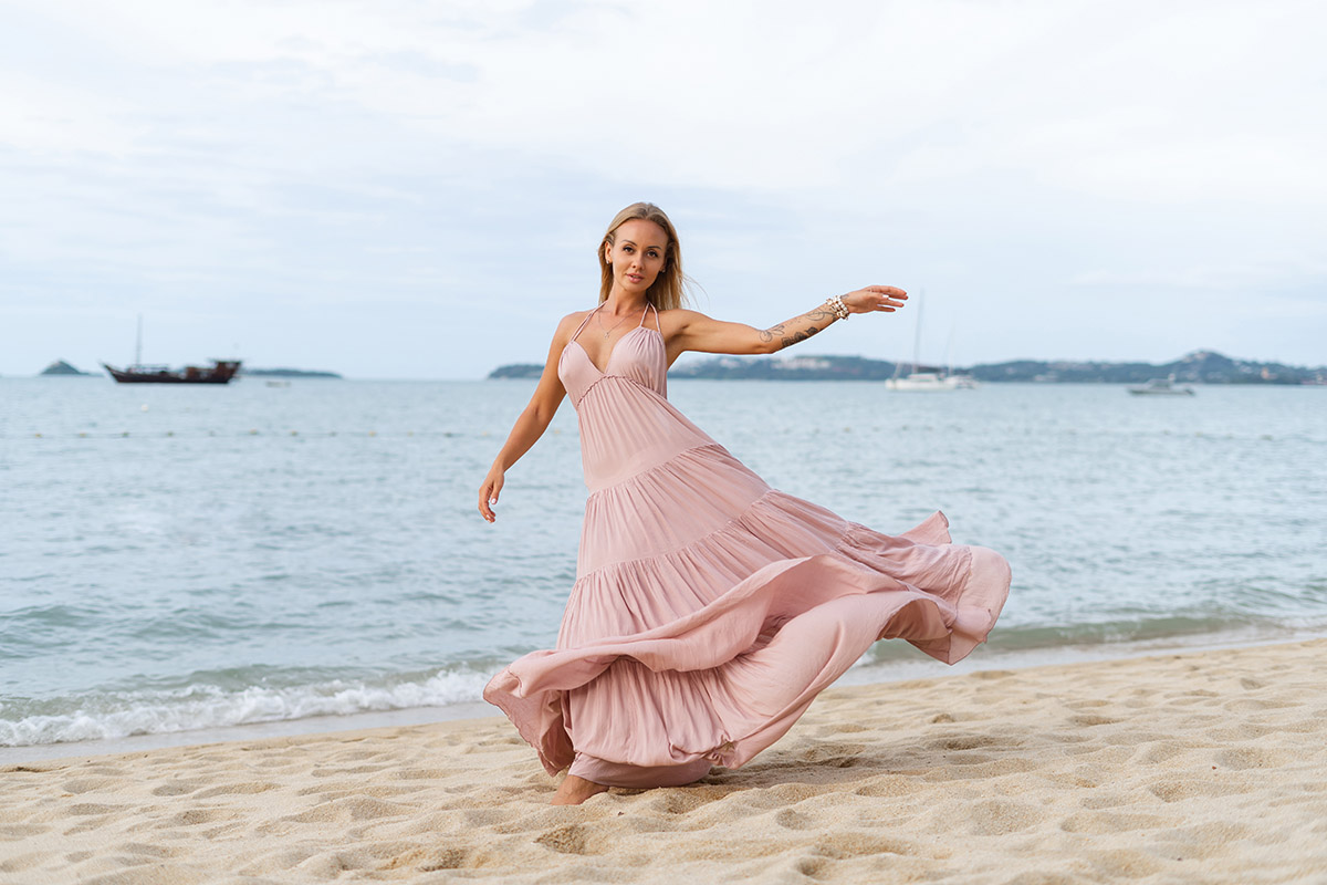 Schöne junge blonde Frau in einem langen rosafarbenen Kleid am Strand, zart, posierend, der Wind entwickelt die Kleiderhaare, romantisch, frei